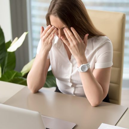 Exhausted businesswoman sitting at desk with laptop. Female office worker suffering from migraine headache during working day. Girl feeling unhealthy at work. Chronic stress and overwork concept